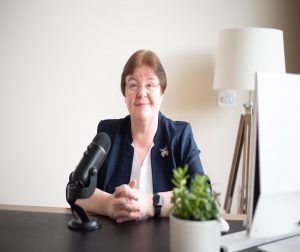 Speaking with Authority. A picture of Anne sitting at a desk. There is a microphone in front of her. She is wearing a navy blue jacket over a white top. She has short brown hair and wears rimless glasses. There is a computer to her right and a lamp in the background.